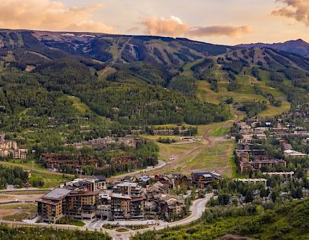 The image shows a scenic mountain resort with buildings nestled among green hills and mountains in the background, under a sky with a mix of clouds and sunset colors.