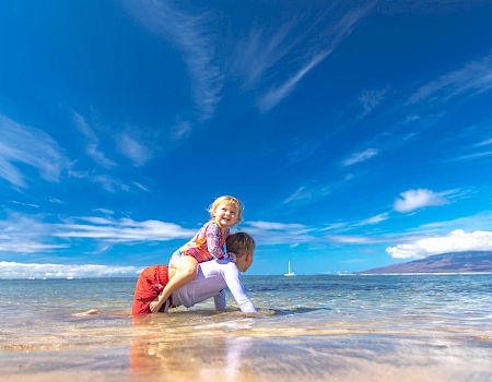 A child is piggybacking on an adult in shallow ocean water under a clear blue sky with some clouds, creating a joyful and playful scene.