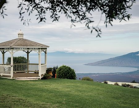 A white gazebo stands on a grassy hill, overlooking a scenic view of mountains and a body of water, with leafy trees framing the foreground.