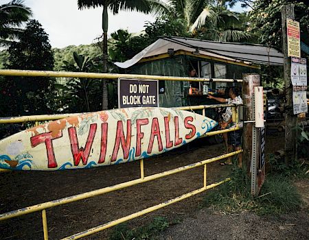 A gate with a sign reading "TWIN FALLS" and another saying "DO NOT BLOCK GATE," with a small building and foliage in the background.