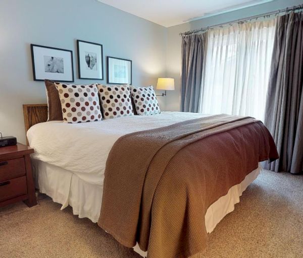 A bedroom with a large bed, polka dot pillows, two nightstands with lamps, wall art, a window with curtains, and a beige armchair.