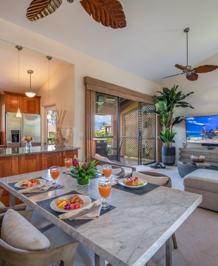 A modern open-concept living area with a kitchen, dining table set with fruit dishes, a cozy lounge, a large TV, tropical decor, and indoor plants.