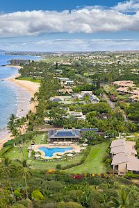 A lush coastal resort town with a sandy beach, blue ocean, palm trees, and clusters of villas and pools, set along a sunny shoreline.