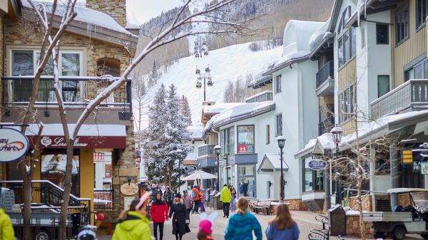 A snow-covered street scene with people walking, surrounded by shops and buildings, and a ski lift visible in the background.