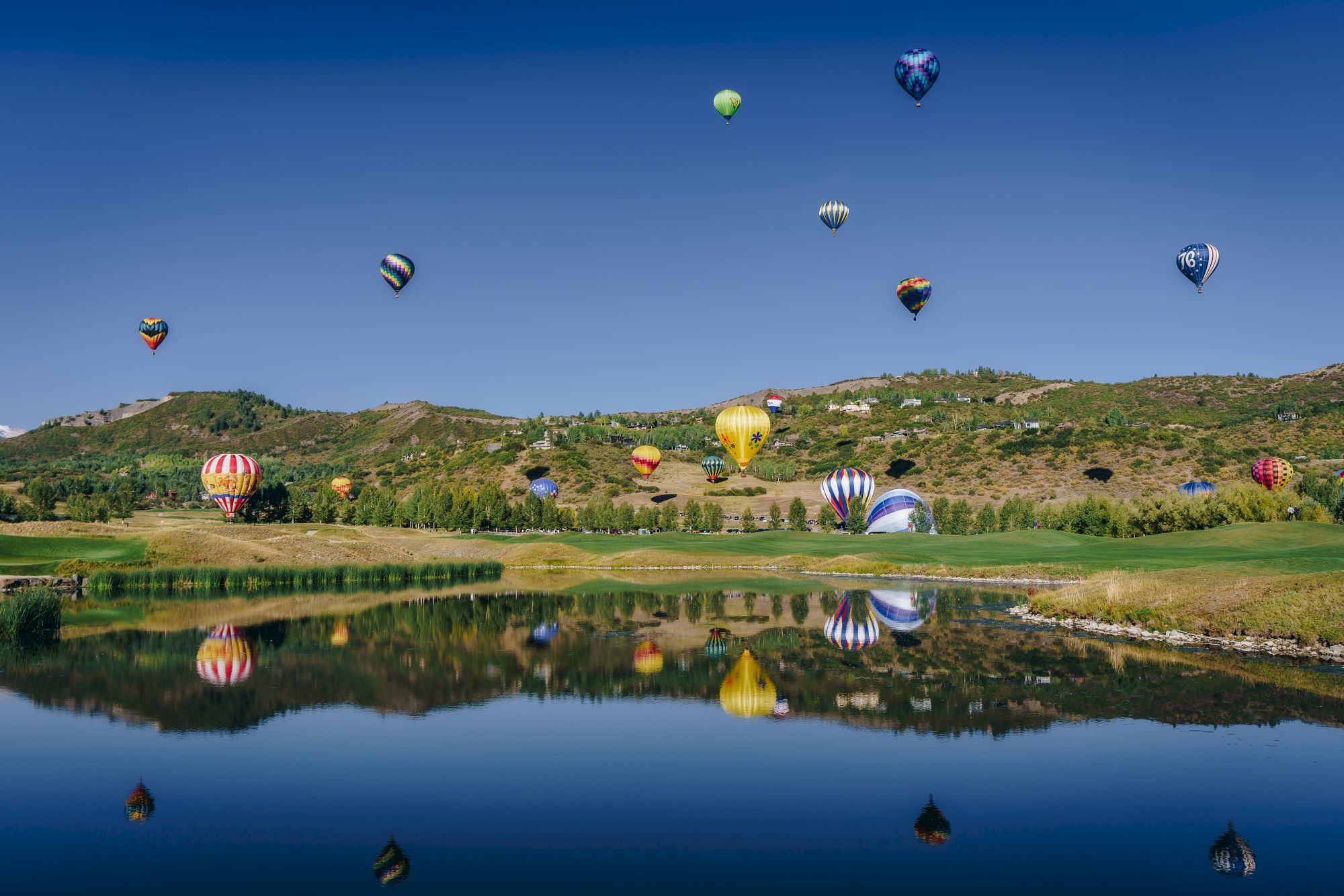 A scenic landscape shows hot air balloons floating above a green hill with a clear blue sky, reflected in a still lake.