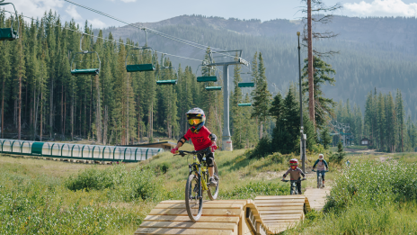 Three cyclists ride on a wooden trail in a scenic forested area with ski lifts visible in the background under a clear blue sky.