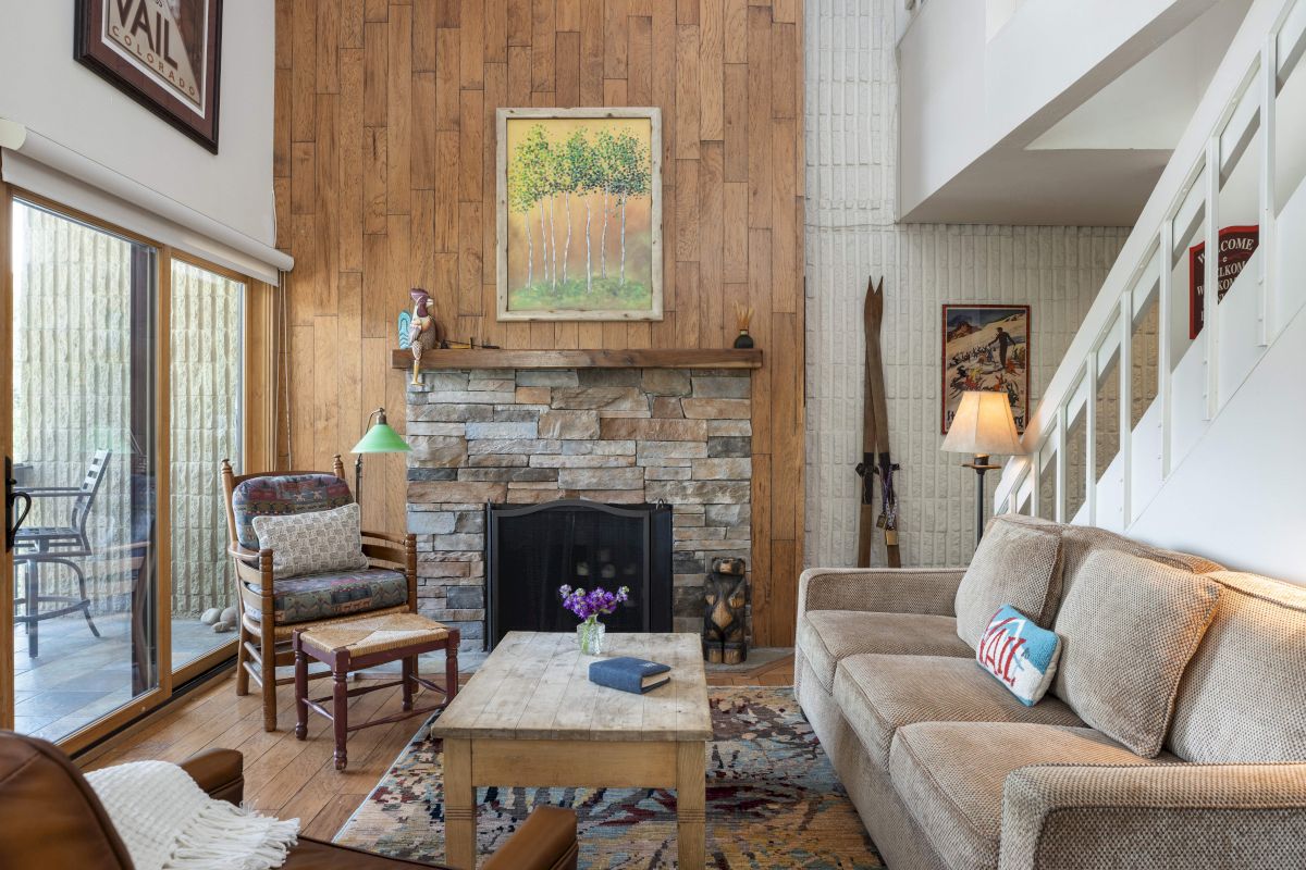 A cozy living room with a stone fireplace, wooden paneling, sofa, chair, coffee table, artwork, and a sliding glass door to a patio.