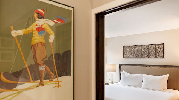 A vintage skiing poster is on the wall near a cozy bedroom with a neatly made bed and a lamp in the corner, creating a warm ambiance.
