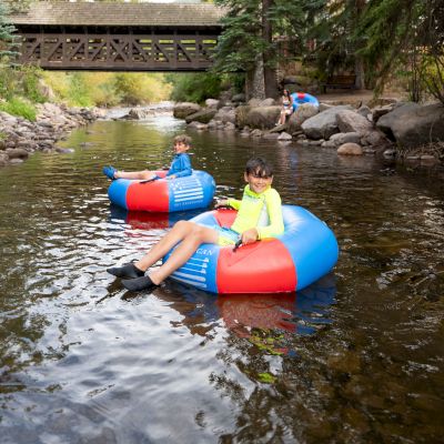 People are floating on colorful tubes in a shallow river, surrounded by trees and rocks, with a wooden bridge in the background.