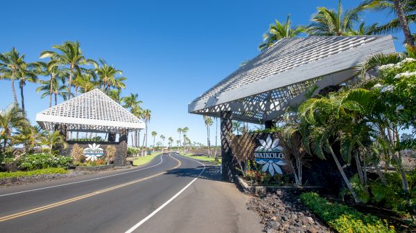 The image shows entrance gates to Waikoloa, surrounded by palm trees and clear blue skies, with a winding road ahead.